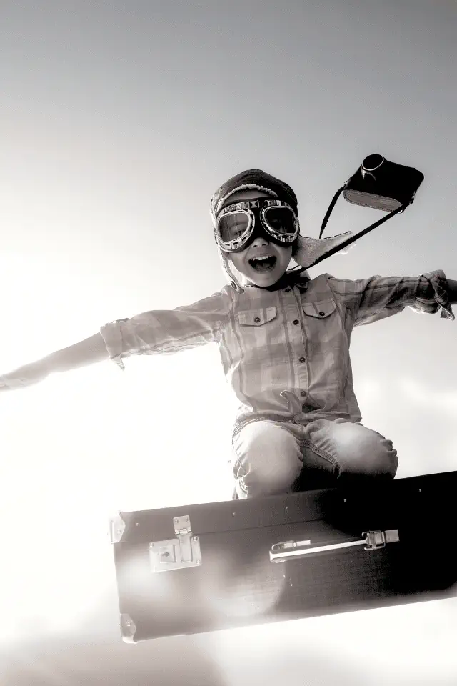 Bambino che sogna di volare, indossando occhiali da aviatore e sorridendo mentre si libra su una valigia. Sognare fa bene a tutte le età.