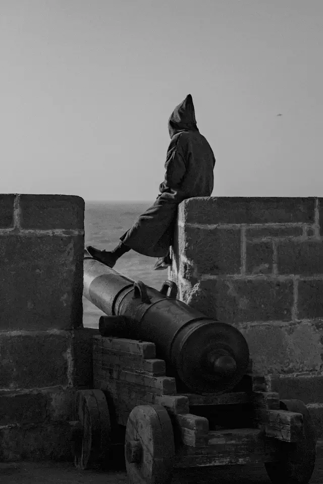 Uomo in silenzio seduto accanto a un cannone antico: metafora della guerra quotidiana combattuta con le parole.