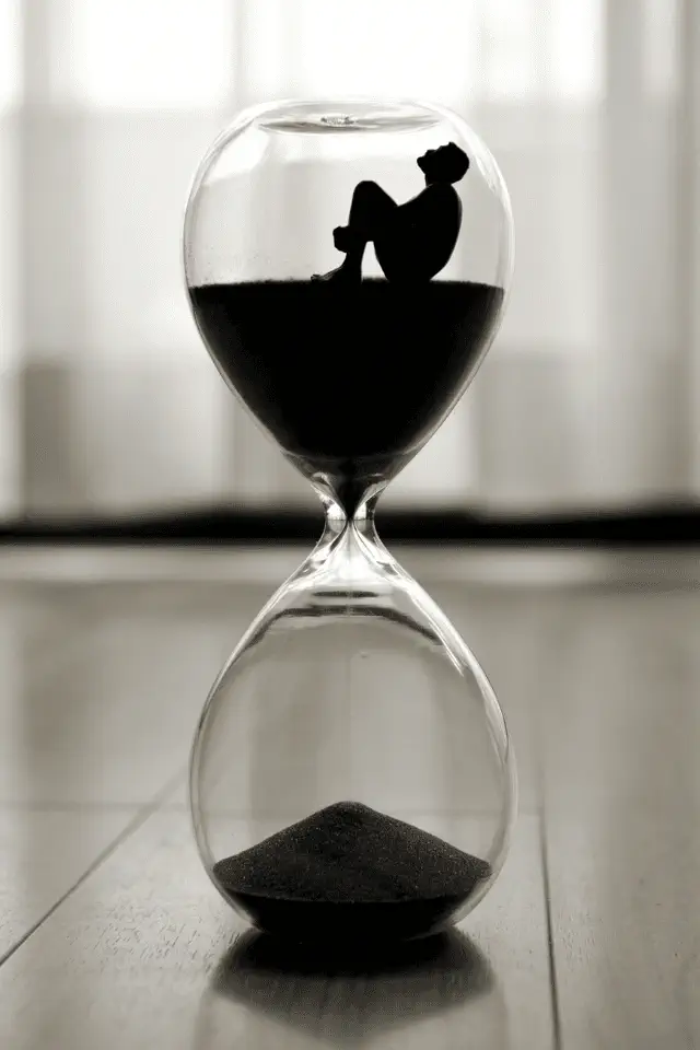 Sagoma di una persona seduta nella parte superiore di una clessidra, circondata da sabbia nera, simbolo dell'illusione di poter ingannare il tempo, mentre lui continua a scorrere.