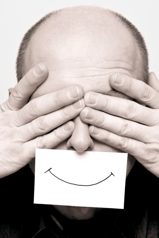 Uomo con occhi coperti e sorriso disegnato su un foglio, simbolo della difficoltà e del disagio nell'essere felici.