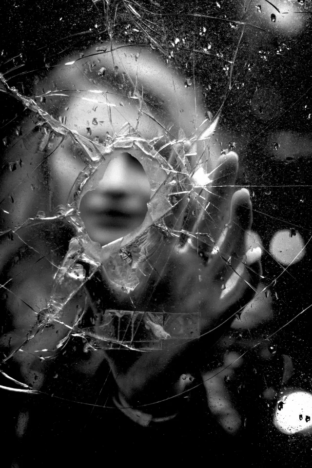 Persona nascosta dietro un vetro rotto con la mano appoggiata, simbolo di vergogna e fragilità umana.